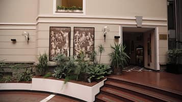 A hotel lobby with a grand staircase, two framed posters, and a variety of indoor plants.