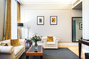 Habitación de hotel con dos sillones blancos, una mesita de centro, un florero con flores y dos cuadros en la pared.
