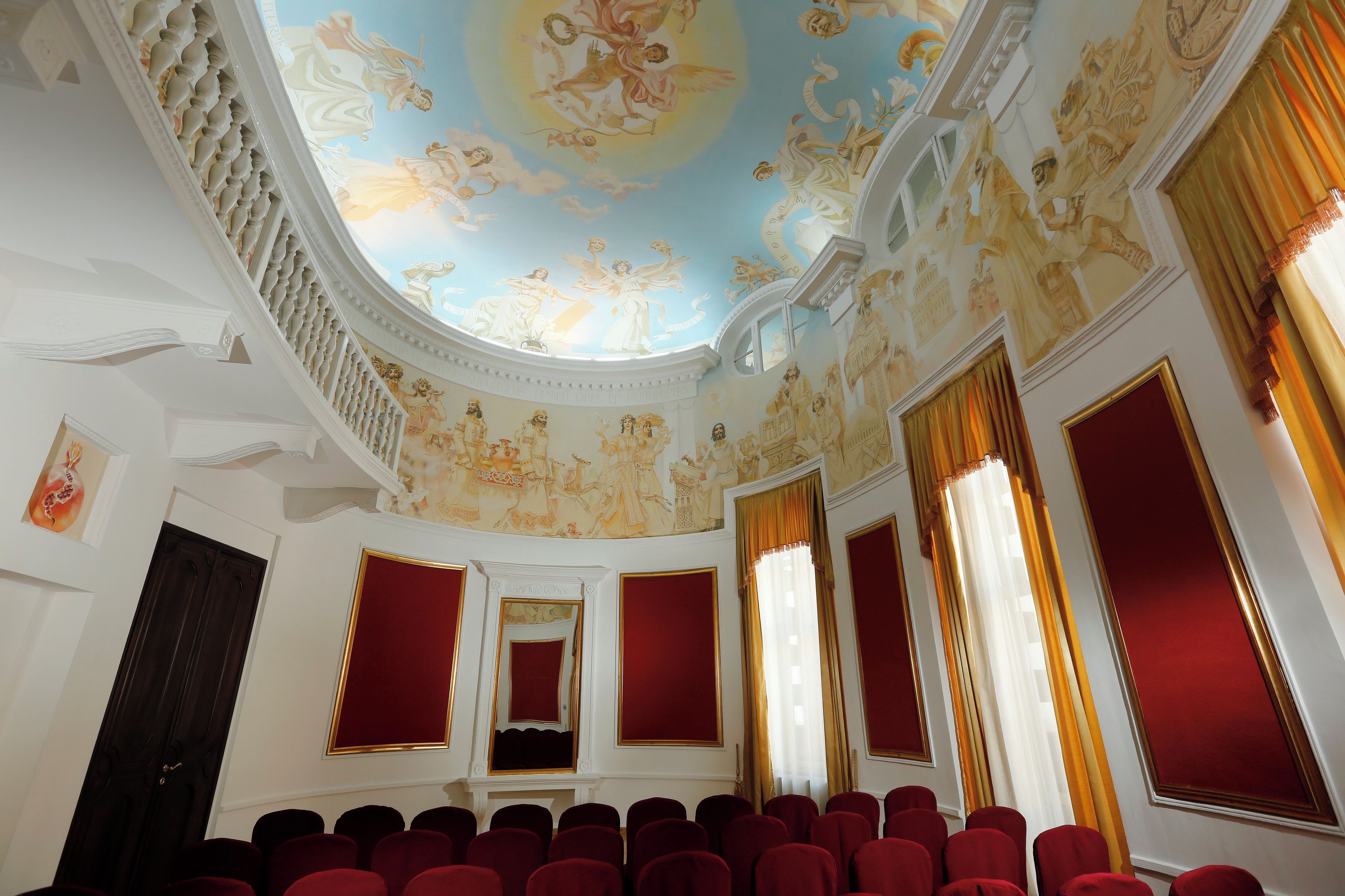 A grand hall with a high ceiling, ornate ceiling artwork, and red upholstered chairs arranged in rows.