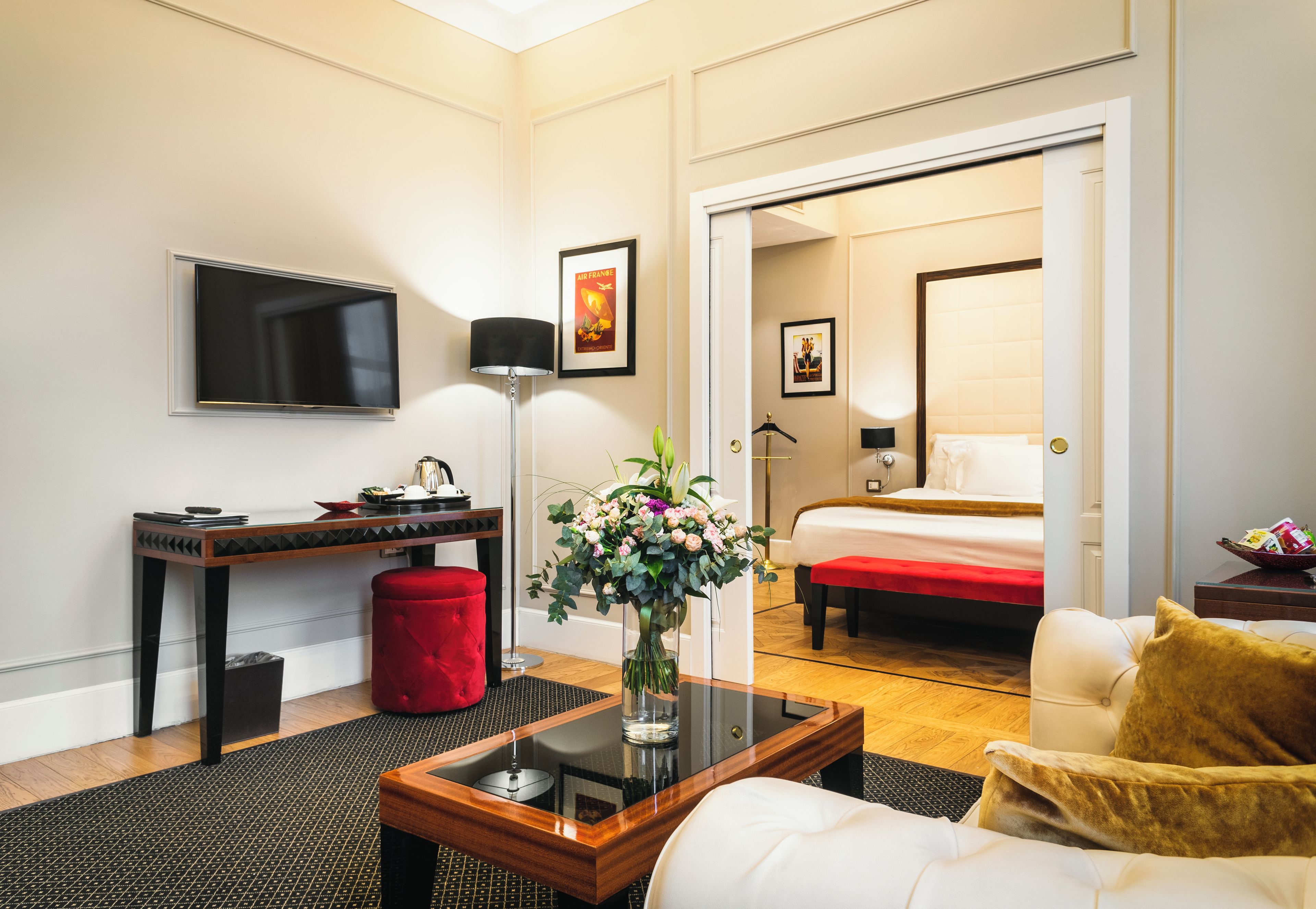 Una habitació d'hotel amb un llit, un escriptori, una televisió, un ram de flors i un otomà vermell.