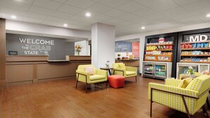 Un vestíbulo de hotel con mostrador de recepción, zona de estar con sillones y reposapiés, y una barra de aperitivos con diversas opciones de comida.