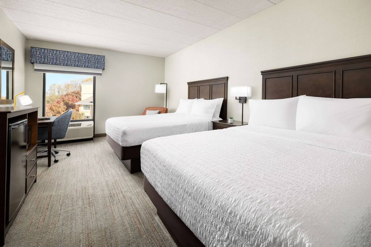 A hotel room with two beds, a desk, a chair, and a large window providing a view of trees.