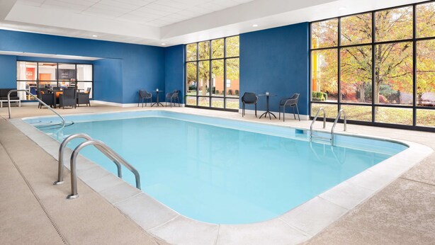 Una piscina con barandillas metálicas, un área de descanso con sillas y grandes ventanales con vistas a árboles.