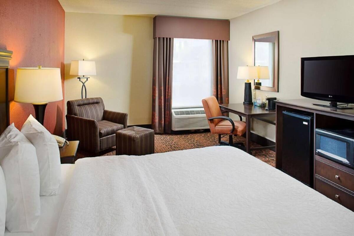 A hotel room with a bed, two armchairs, a desk with a television, a mirror, and an air conditioning unit.