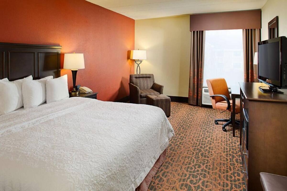 A hotel room with a large bed, a desk with a television, a chair, and a lamp.