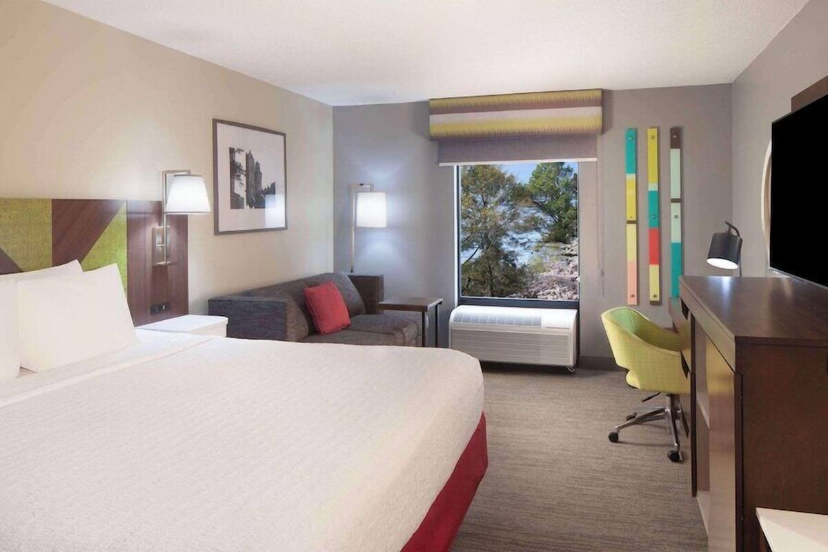 A hotel room with a large bed, a desk, a chair, a TV, and a window with a view of trees.