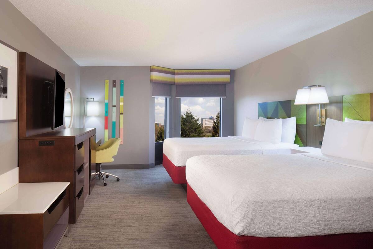 A hotel room with two beds, a desk, a TV, a window with a view, and a colorful wall decoration.