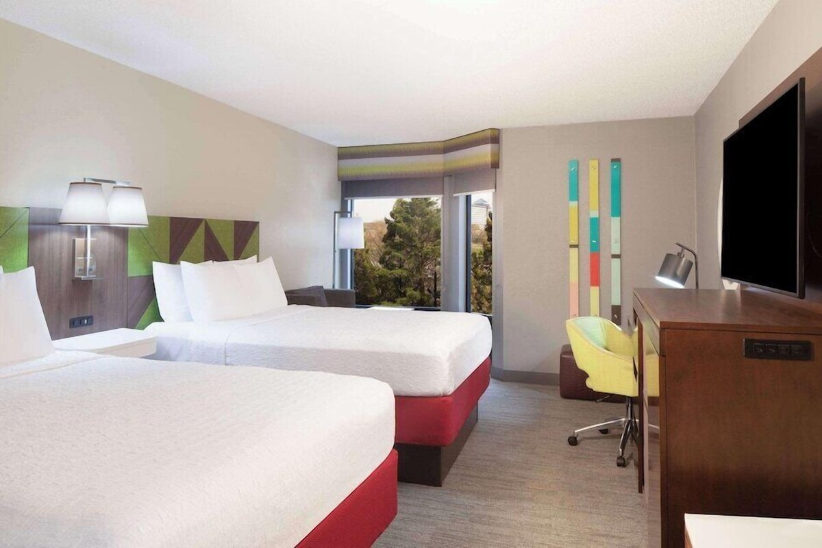A hotel room with two beds, a desk with a chair, a TV, and a window with a view of trees.