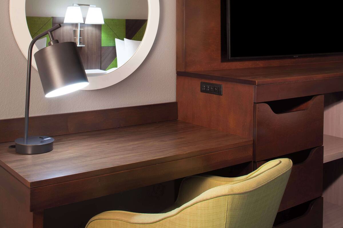 A modern hotel room with a wooden desk, a round mirror, a lamp, a television, and a chair.