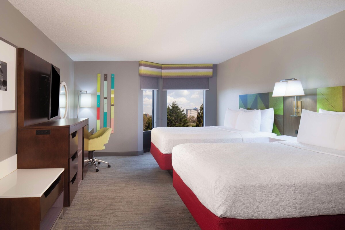 A hotel room with two beds, a desk, a TV, a window with a view, and a colorful wall decoration.