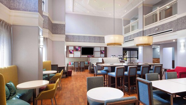 A spacious hotel lobby with multiple seating areas, including round tables and booths, and a large television screen. - Hampton Inn & Suites Atlanta Airport North I-85 (Atlanta)