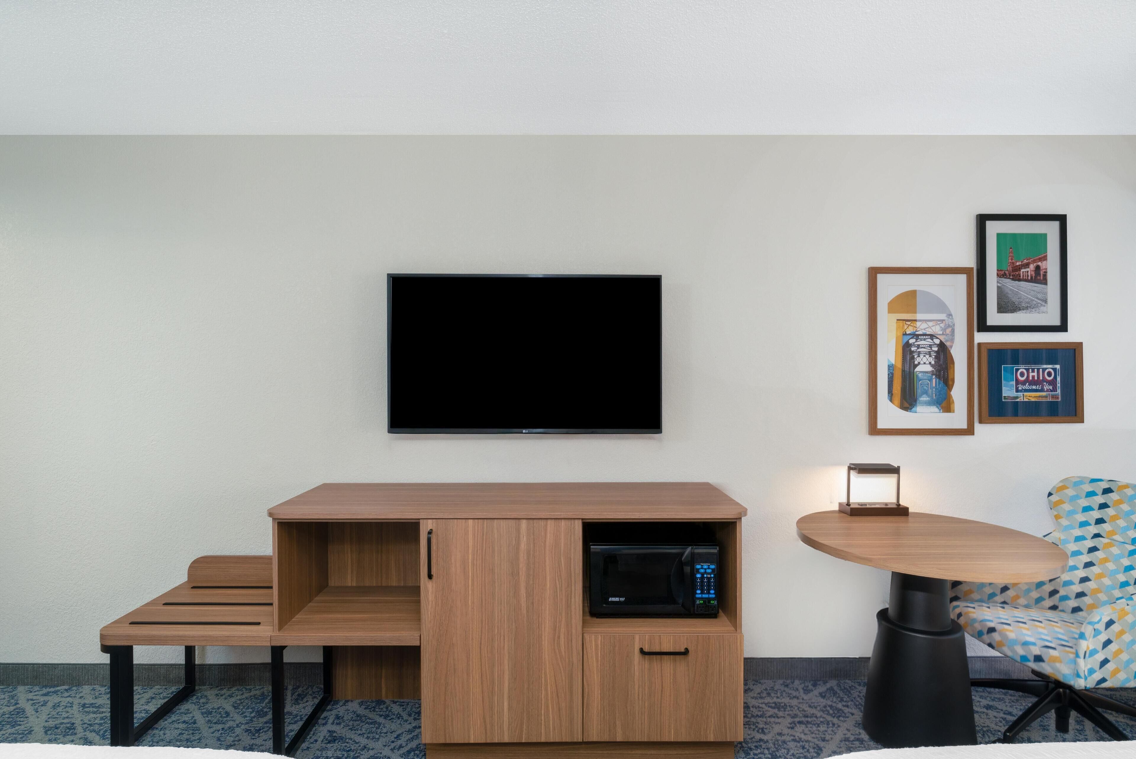 Una habitación de hotel moderna con un televisor de pantalla plana montado en la pared, un mueble de madera con microondas, una mesita con lámpara y un sillón con estampado.