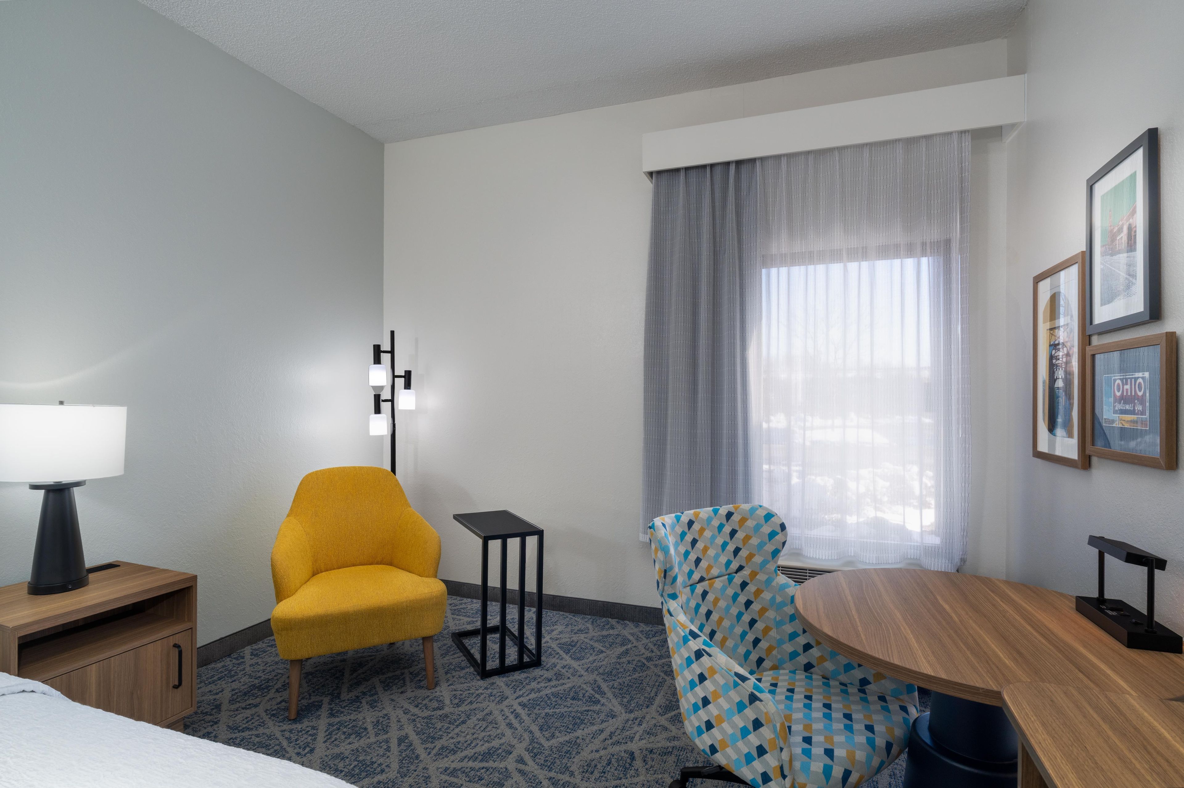 A hotel room with a bed, a yellow armchair, a blue patterned armchair, a small table, a lamp, and a window with curtains.