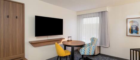 Habitación de hotel con cama, televisor montado en la pared, armario de madera, escritorio con silla y ventana con cortinas.