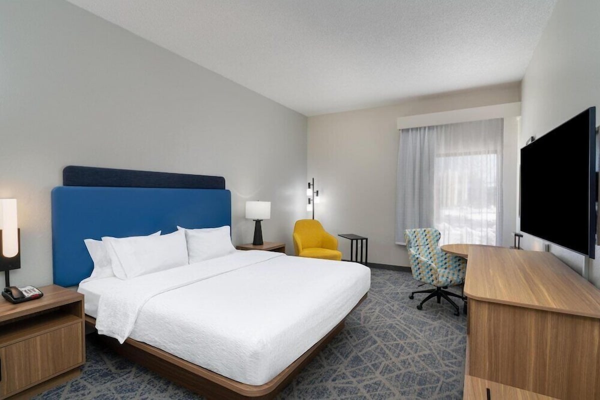 A hotel room with a bed, a desk, a chair, a TV, and a lamp.