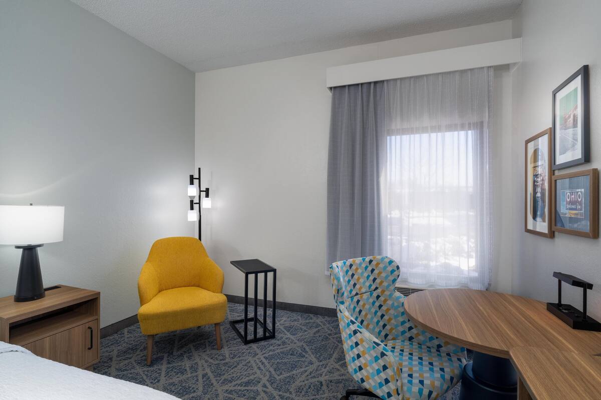 A hotel room with a bed, a yellow armchair, a blue patterned armchair, a small table, a lamp, and a window with curtains.
