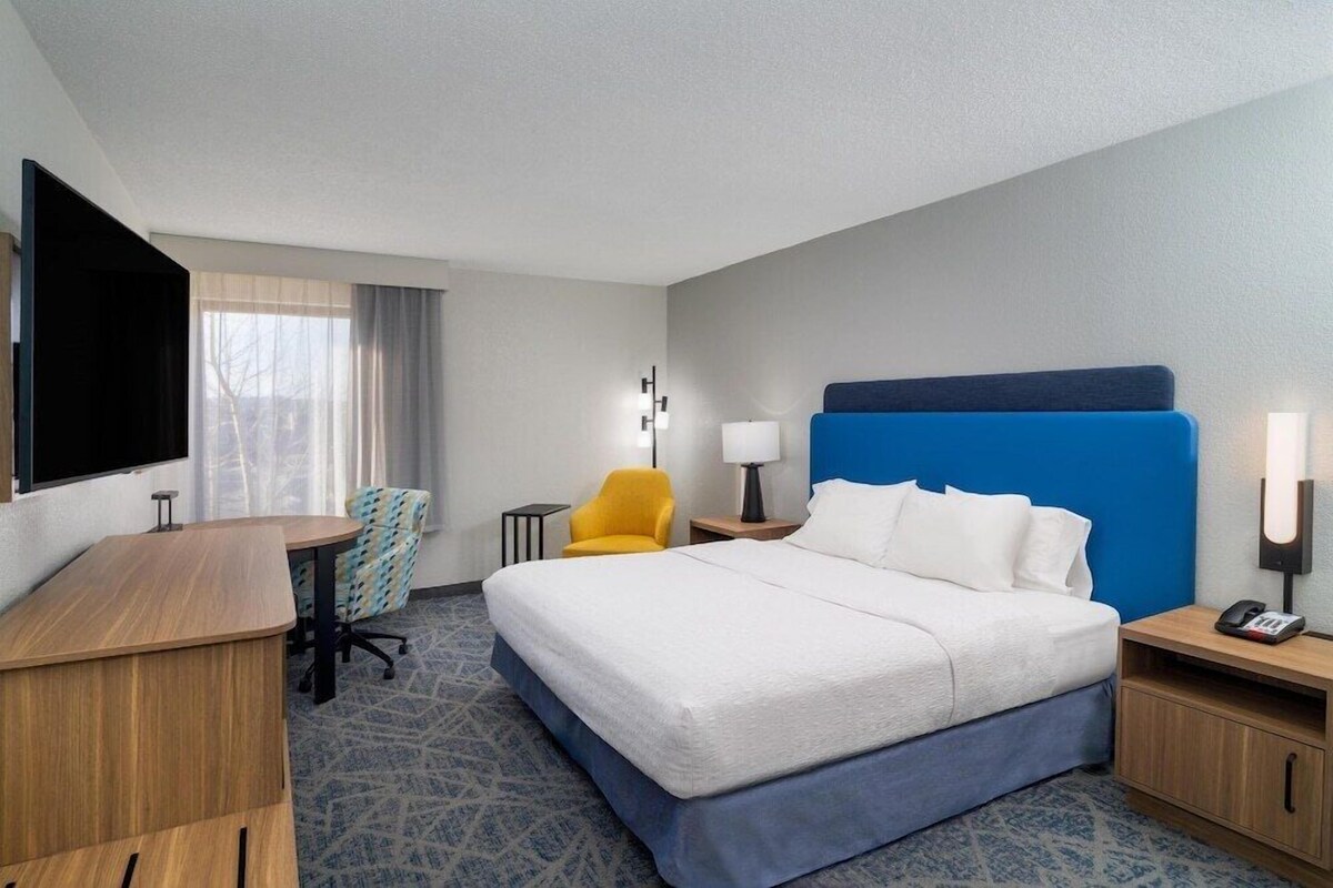 A hotel room with a bed, a desk, a chair, a TV, a lamp, and a nightstand.