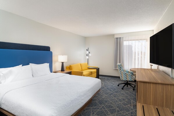 Quarto de hotel com uma cama grande, escrivaninha, cadeira, televisão e janela com cortinas.