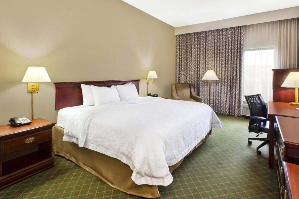 A hotel room with a large bed, two bedside tables with lamps, a desk with a chair, and a patterned curtain.