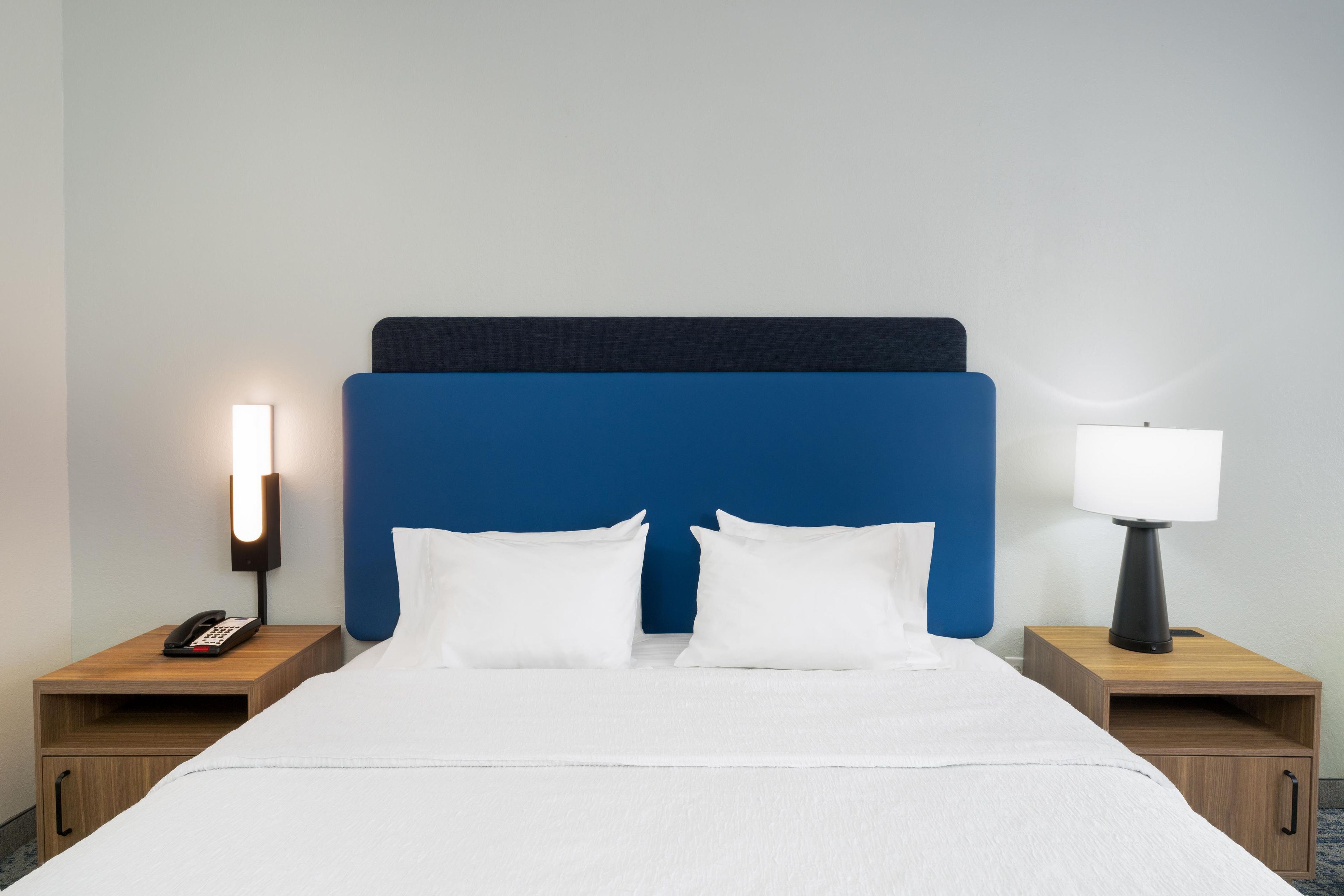 A hotel room with a bed, two bedside tables, a nightstand, a lamp, and a phone.