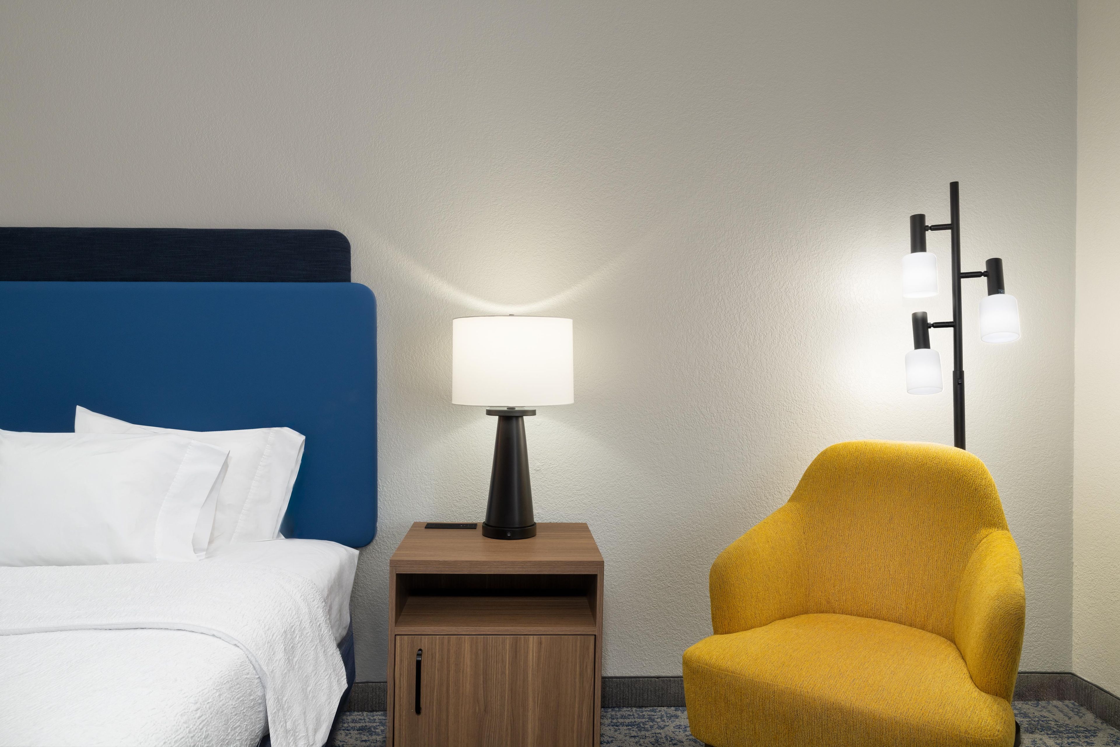 A hotel room with a bed, a bedside table with a lamp, a small wooden cabinet, and a yellow armchair.