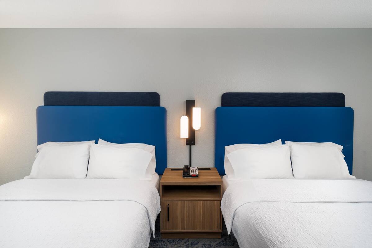 A hotel room with two beds, each with white bedding and blue headboards. There is a nightstand between the beds with a lamp and a telephone.