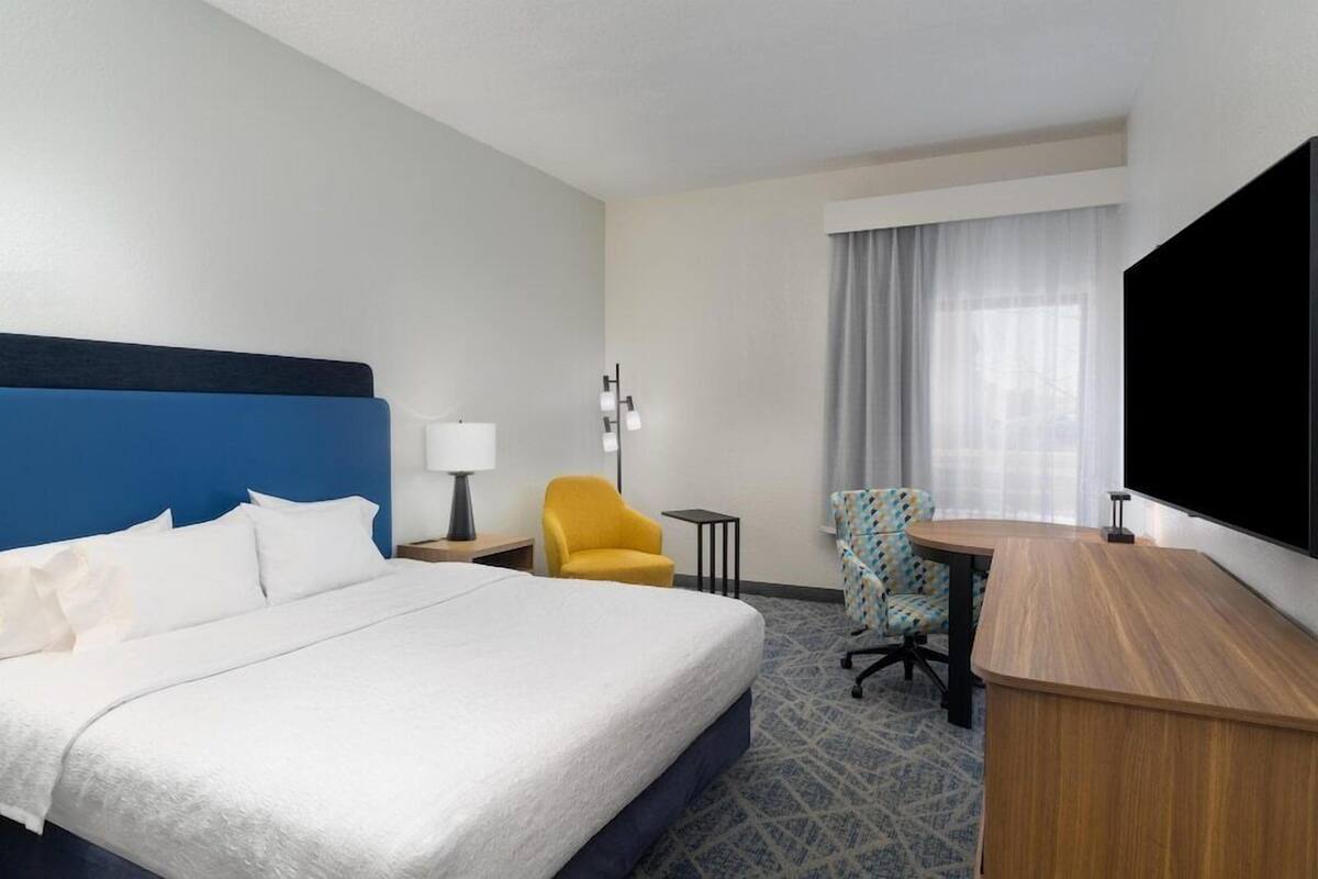 A hotel room with a large bed, a desk, a chair, a lamp, and a TV.