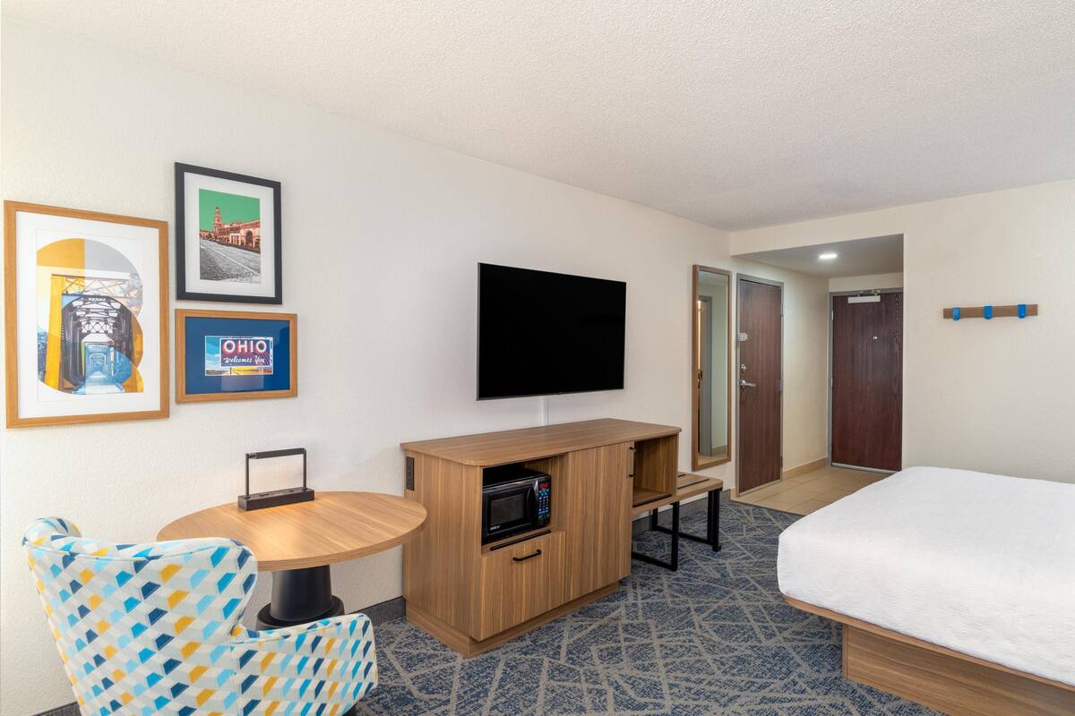 A hotel room with a bed, a TV mounted on the wall, a small wooden table with a microwave, a patterned armchair, and framed pictures on the wall.