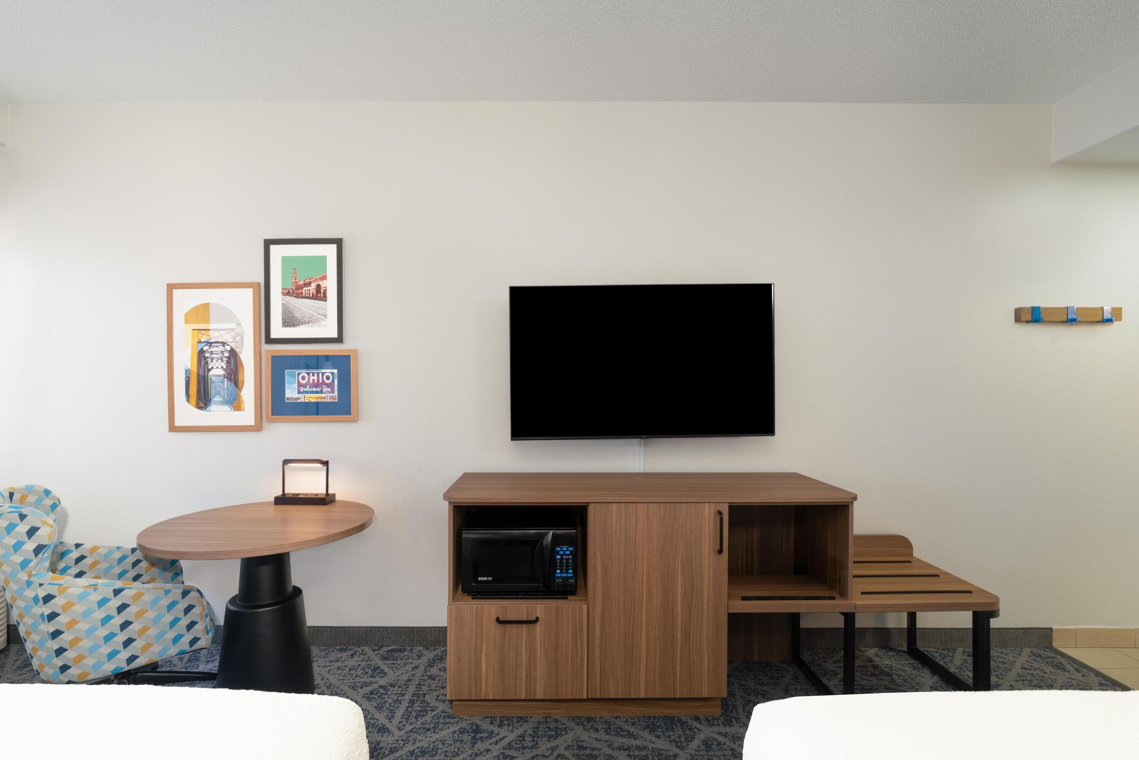 Habitación de hotel con televisor de pantalla plana montado en la pared, mueble de madera con microondas, una mesita y dos sillas estampadas.