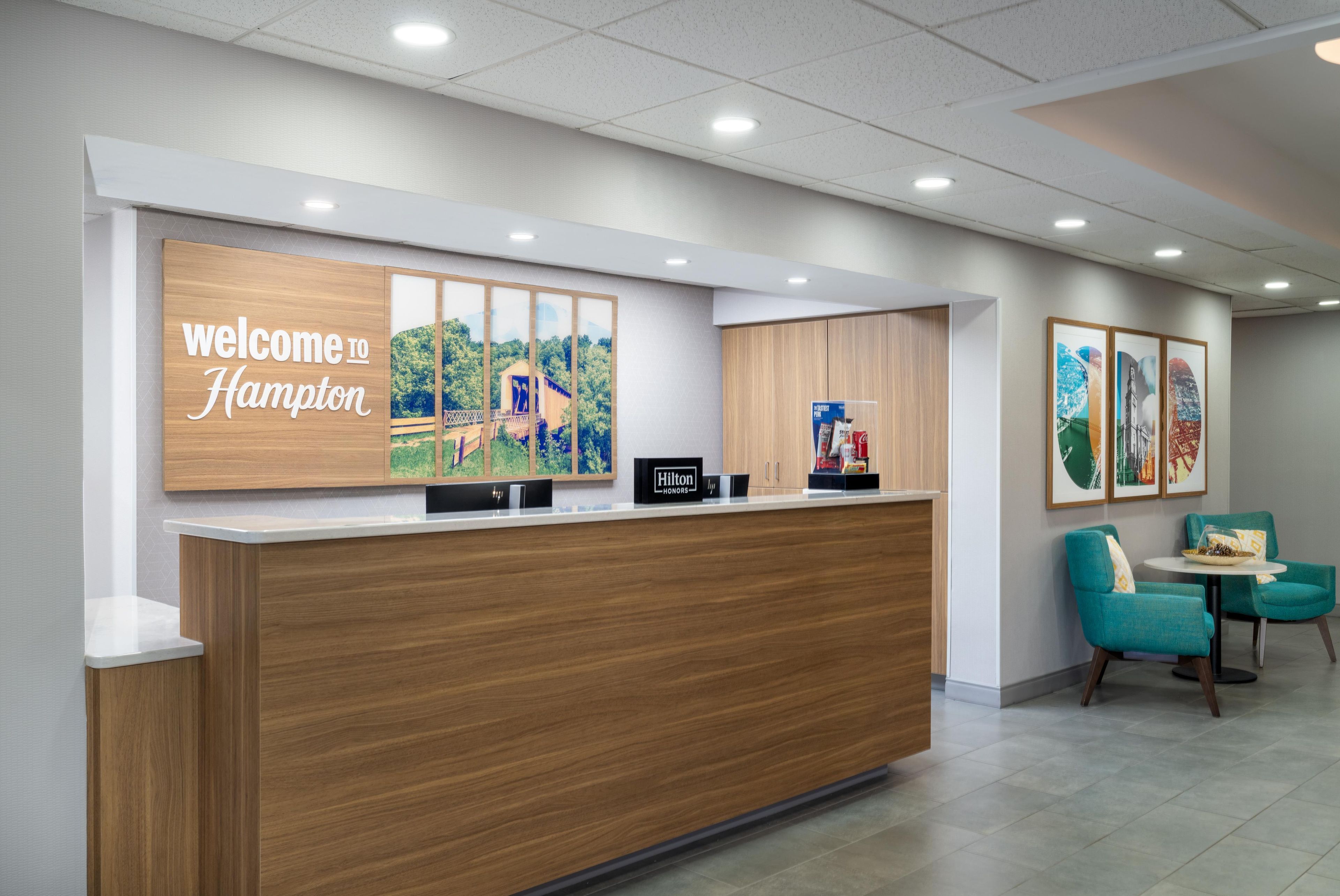 Un comptoir d’accueil avec une pancarte « Bienvenue à Hampton », un tableau représentant un paysage encadré et un coin salon avec des fauteuils de couleur turquoise.