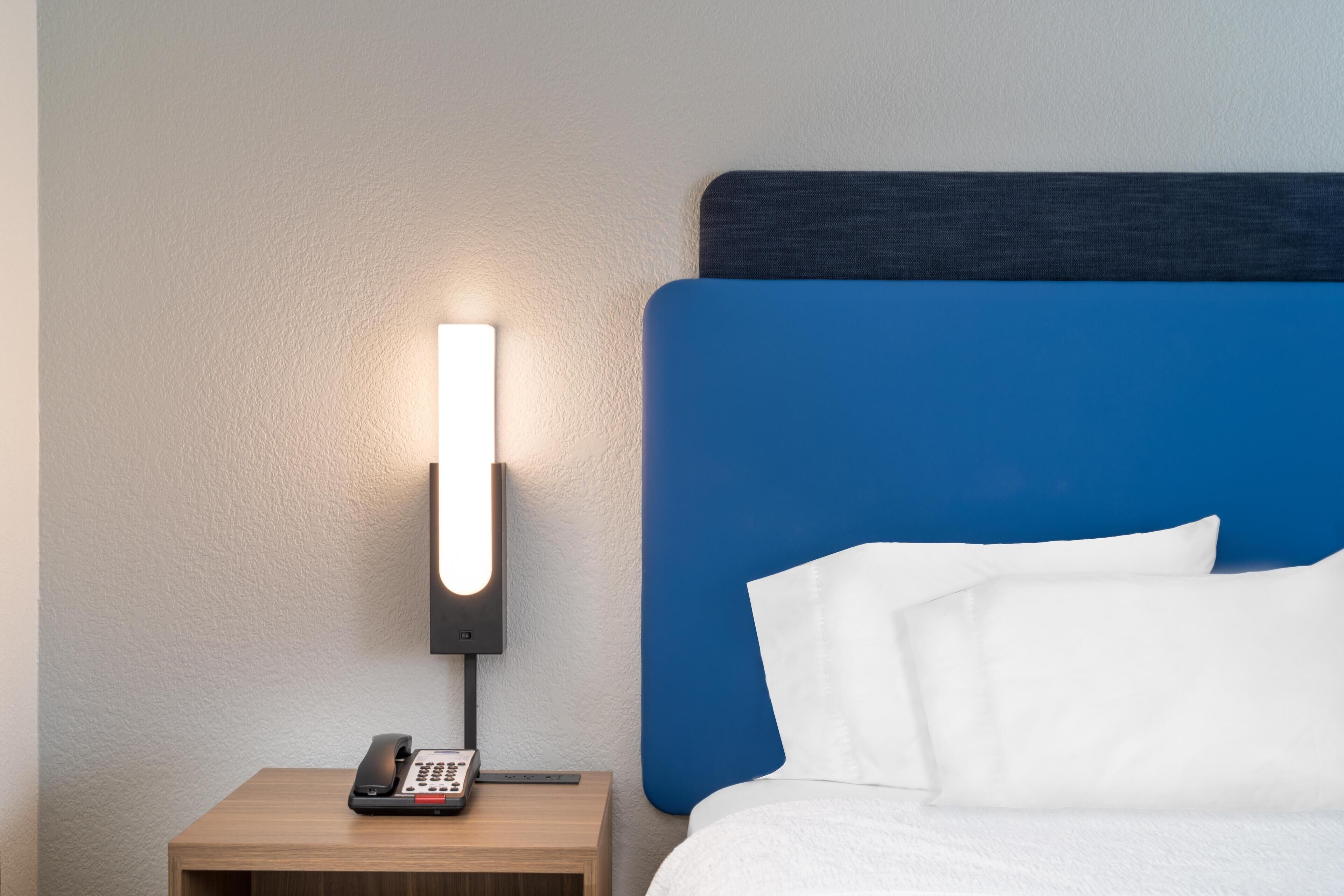 A hotel room with a bed, a nightstand with a phone, and a wall-mounted light.