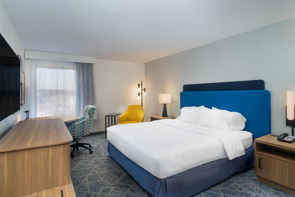 A hotel room with a large bed, a desk, a chair, a nightstand, and a TV.