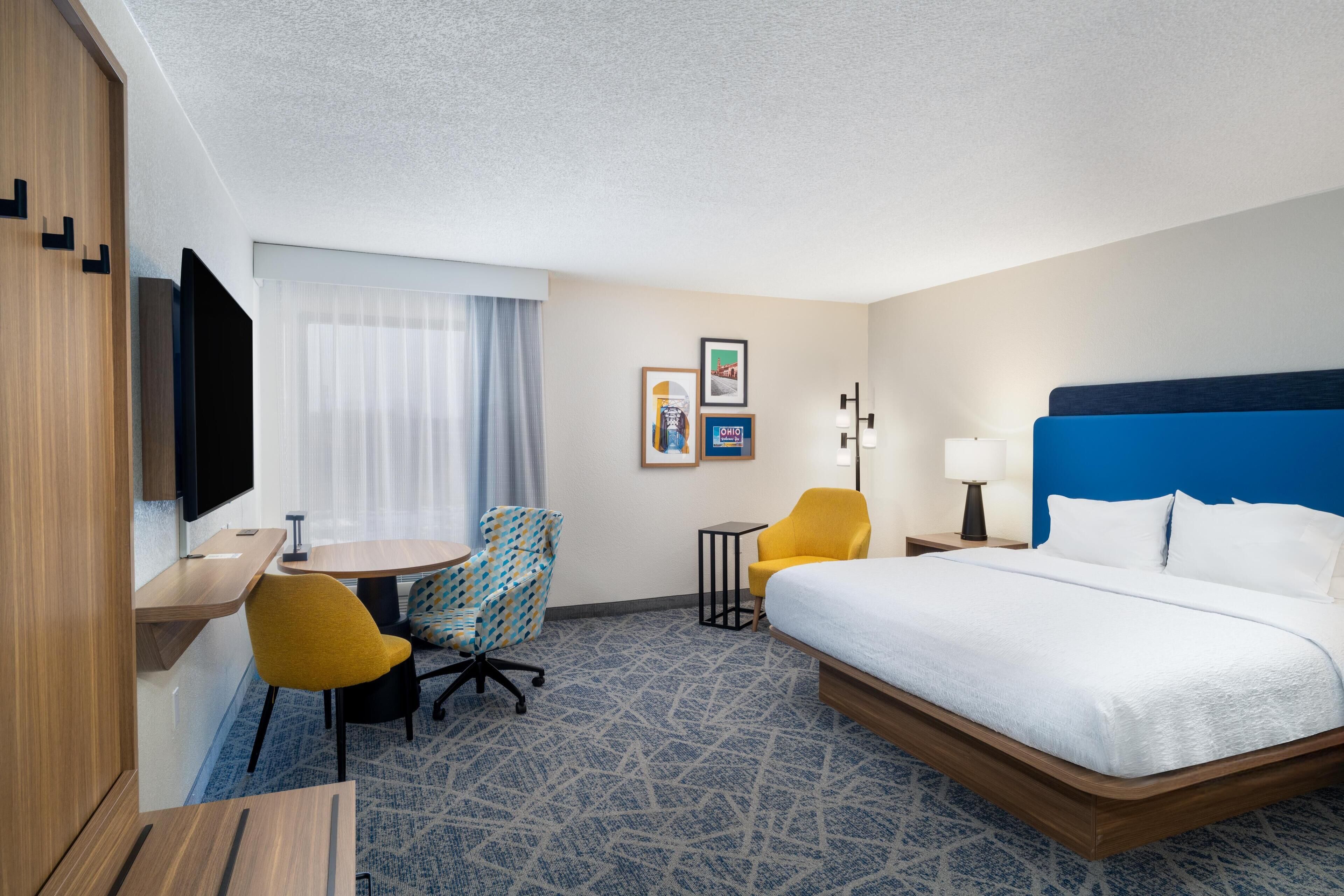 A hotel room with a bed, a desk, a chair, a TV, and a window with curtains.