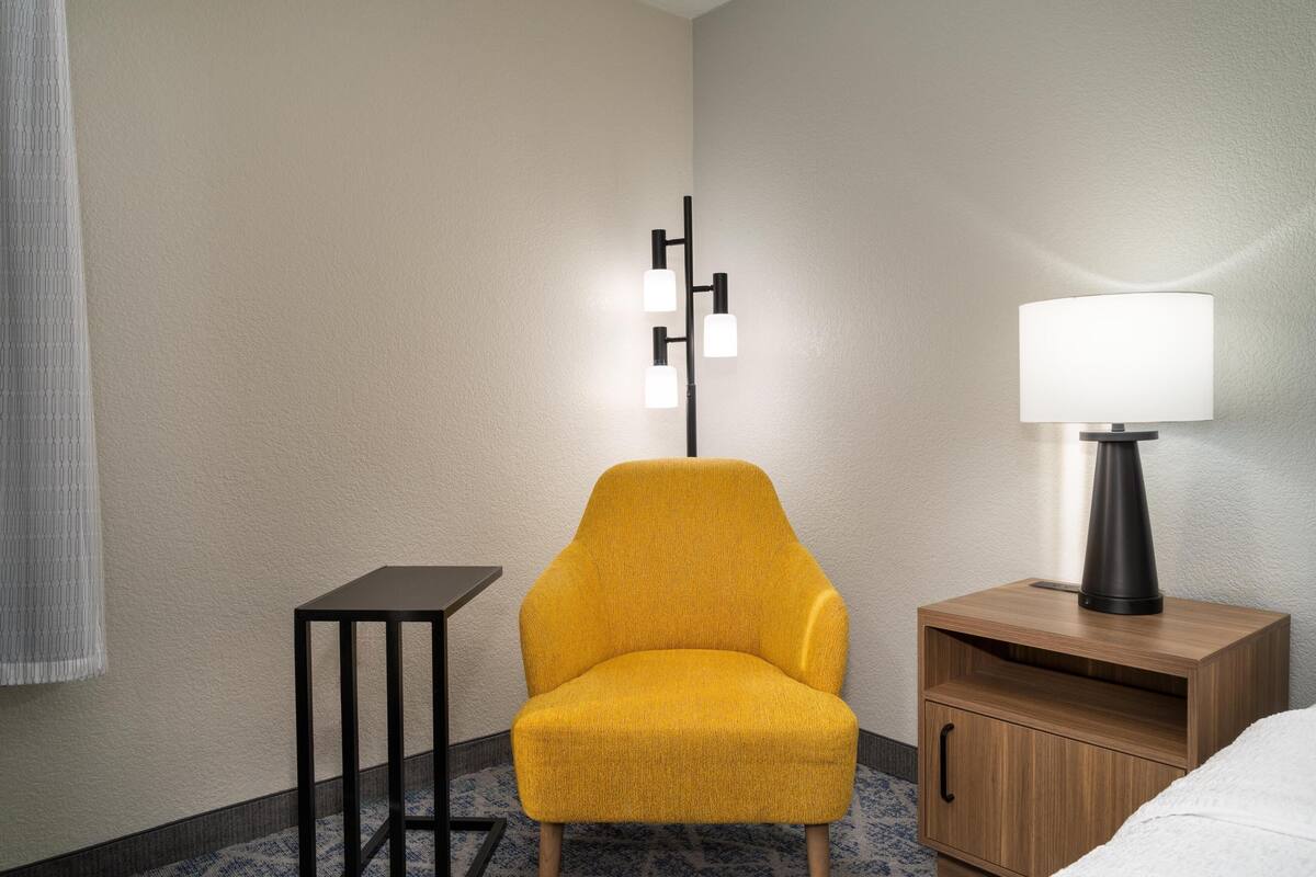 A hotel room with a yellow armchair, a bedside table with a lamp, and a small wooden cabinet.