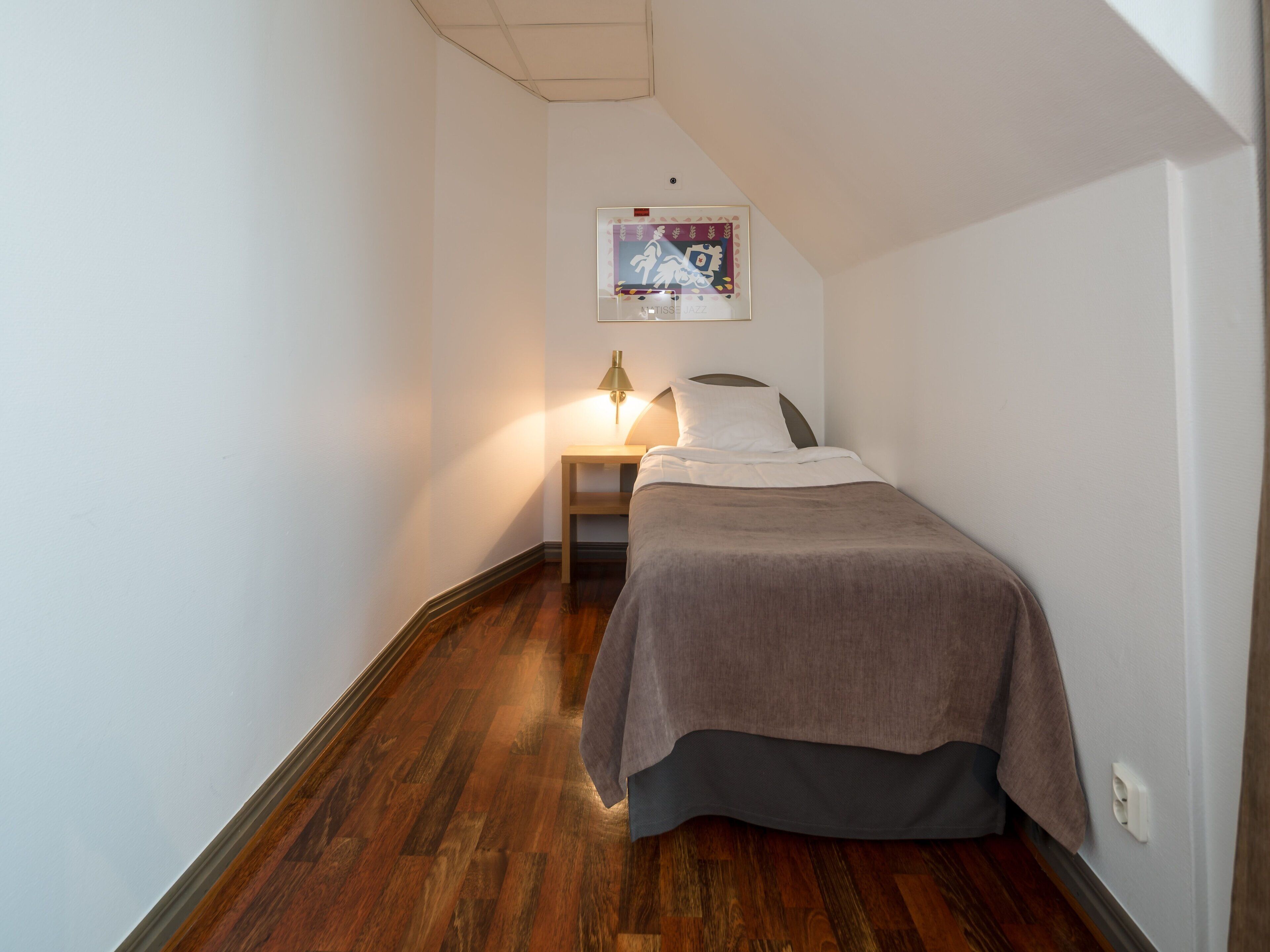 Une petite pièce étroite, avec un lit, une table de chevet et un tableau accroché au mur.