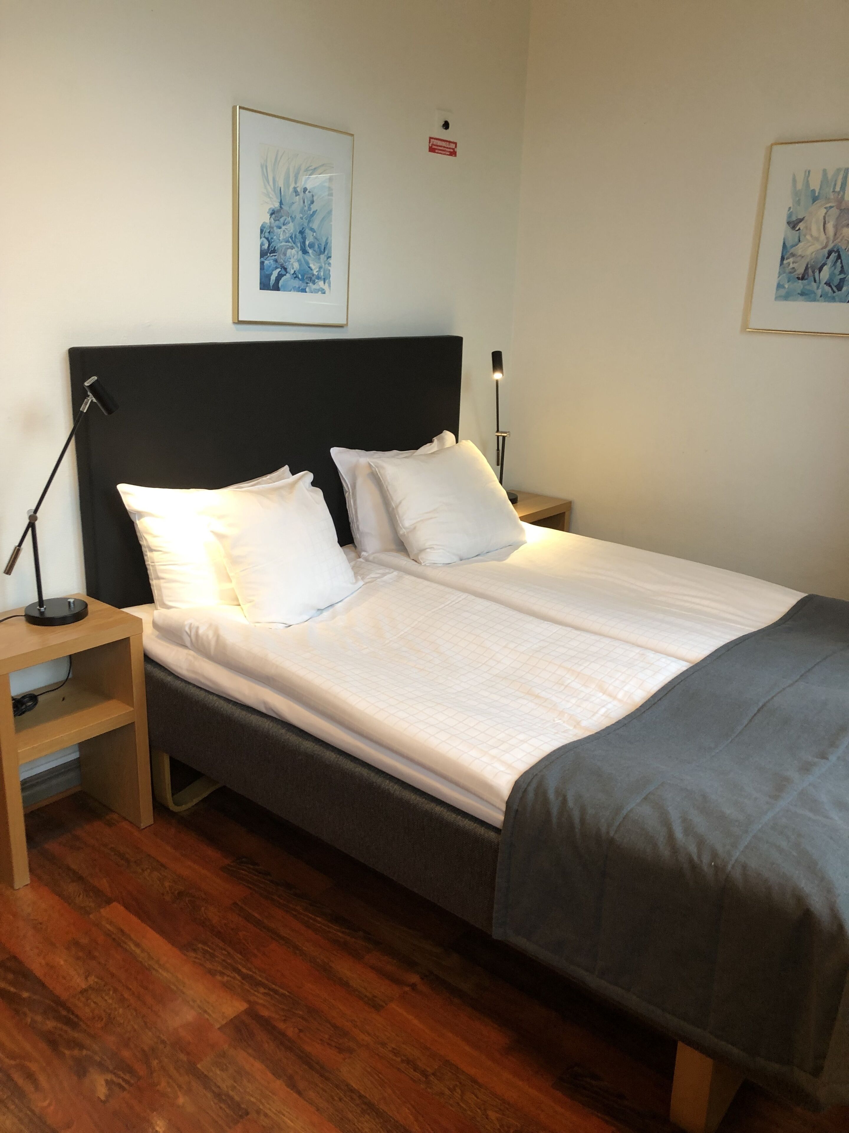 Un lit simple avec une tête de lit noire, du linge de lit blanc et un plaid gris. Il y a deux tables de chevet, l’une avec une lampe et l’autre avec une petite étagère.