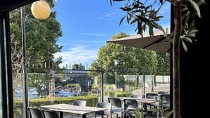 An outdoor patio area with tables and chairs, a view of a river, and a bridge in the background.
