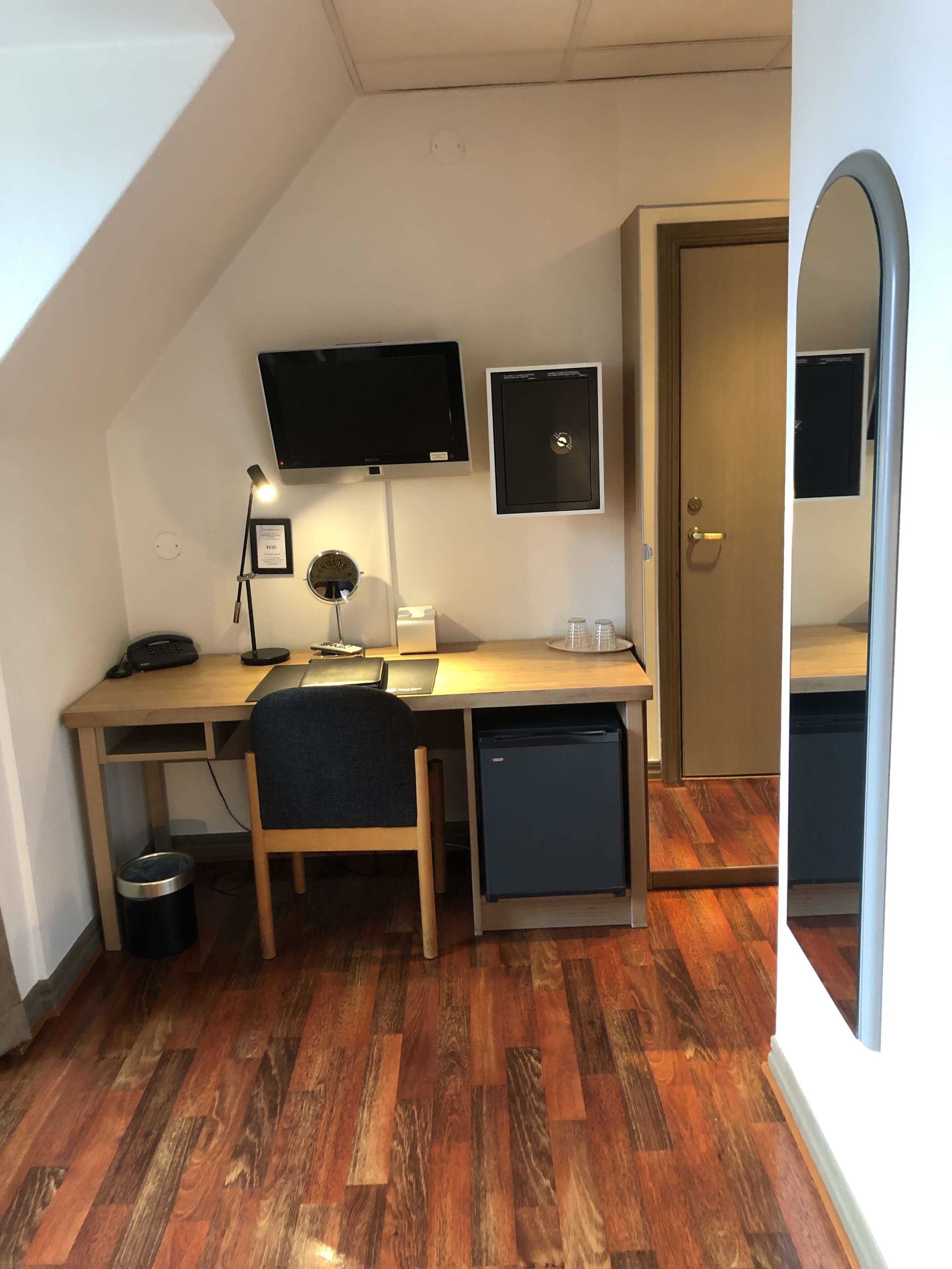 Il s’agit d’une pièce de taille modeste, équipée d’un bureau, d’une chaise et d’un petit réfrigérateur. Un téléviseur est fixé au mur, et une porte donne accès à une autre pièce.