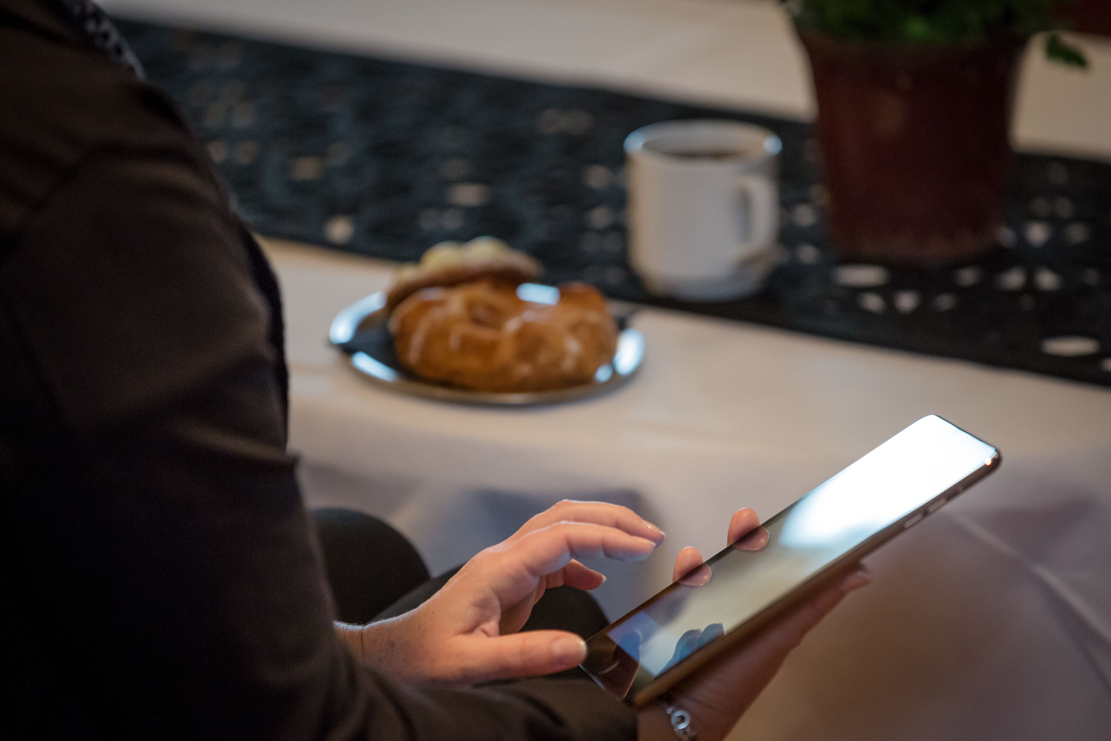 Una persona sosteniendo un teléfono inteligente, con un croissant y una taza sobre una mesa al fondo.