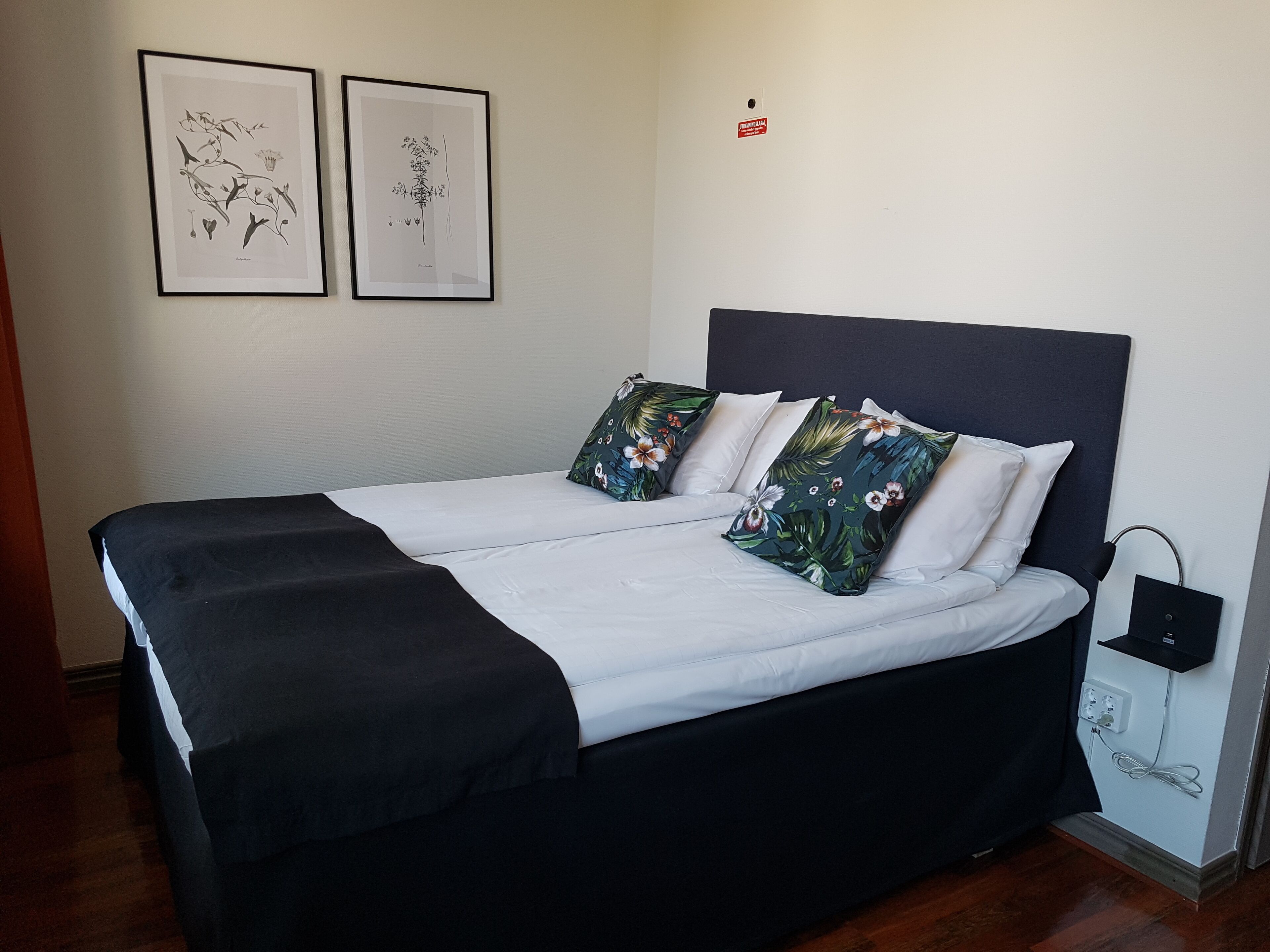 A neatly made bed with a black and white bedspread, two floral pillows, and a dark headboard. There are two framed botanical prints on the wall above the bed.