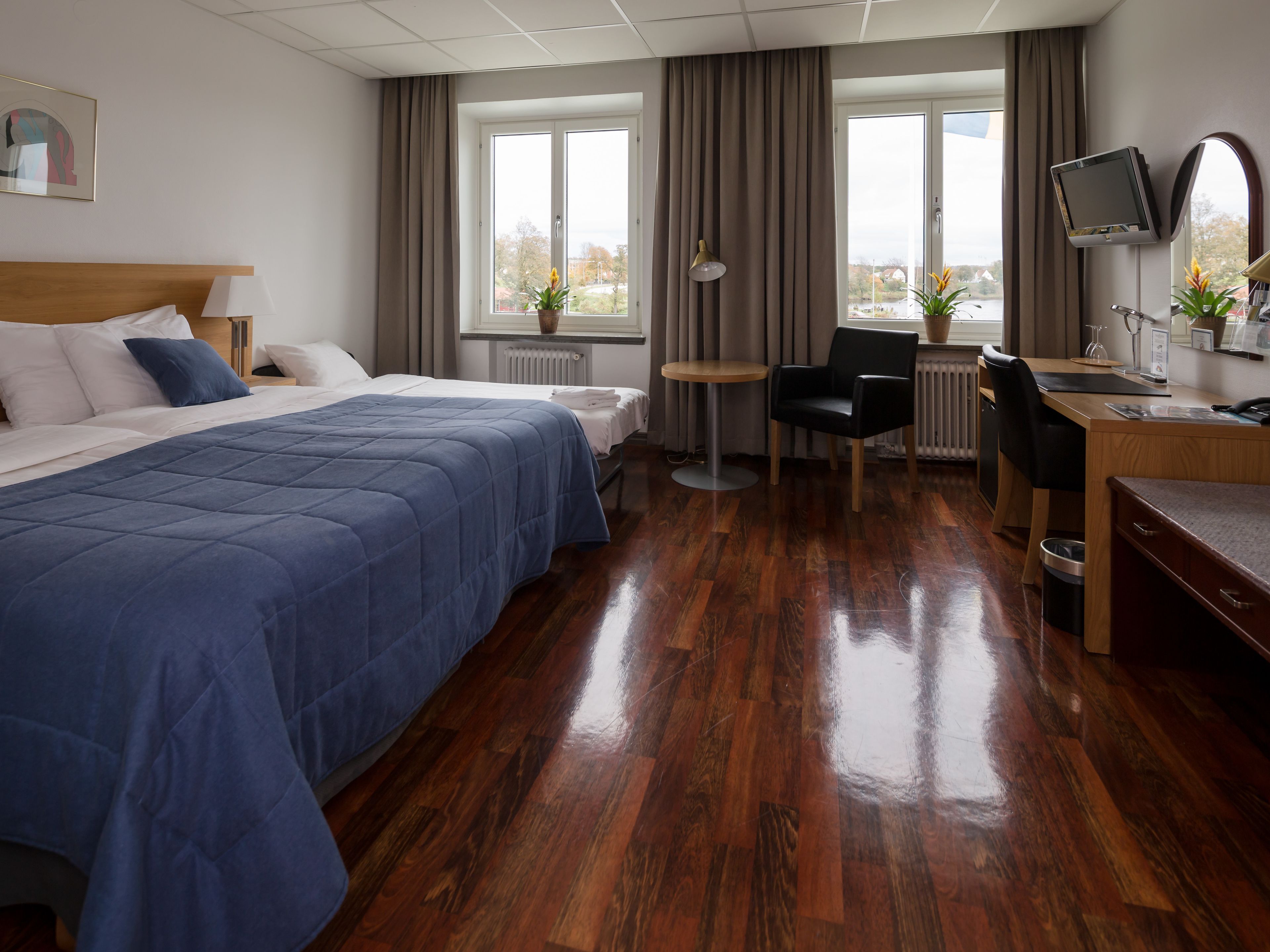 A hotel room with a large bed, a desk, a chair, a small table, a TV, and a window with curtains.