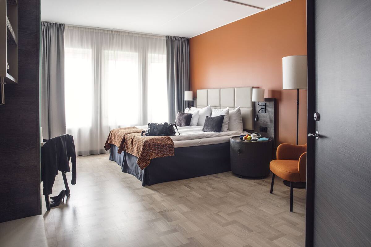 A modern hotel room with a large bed, a desk, a chair, and a wardrobe.