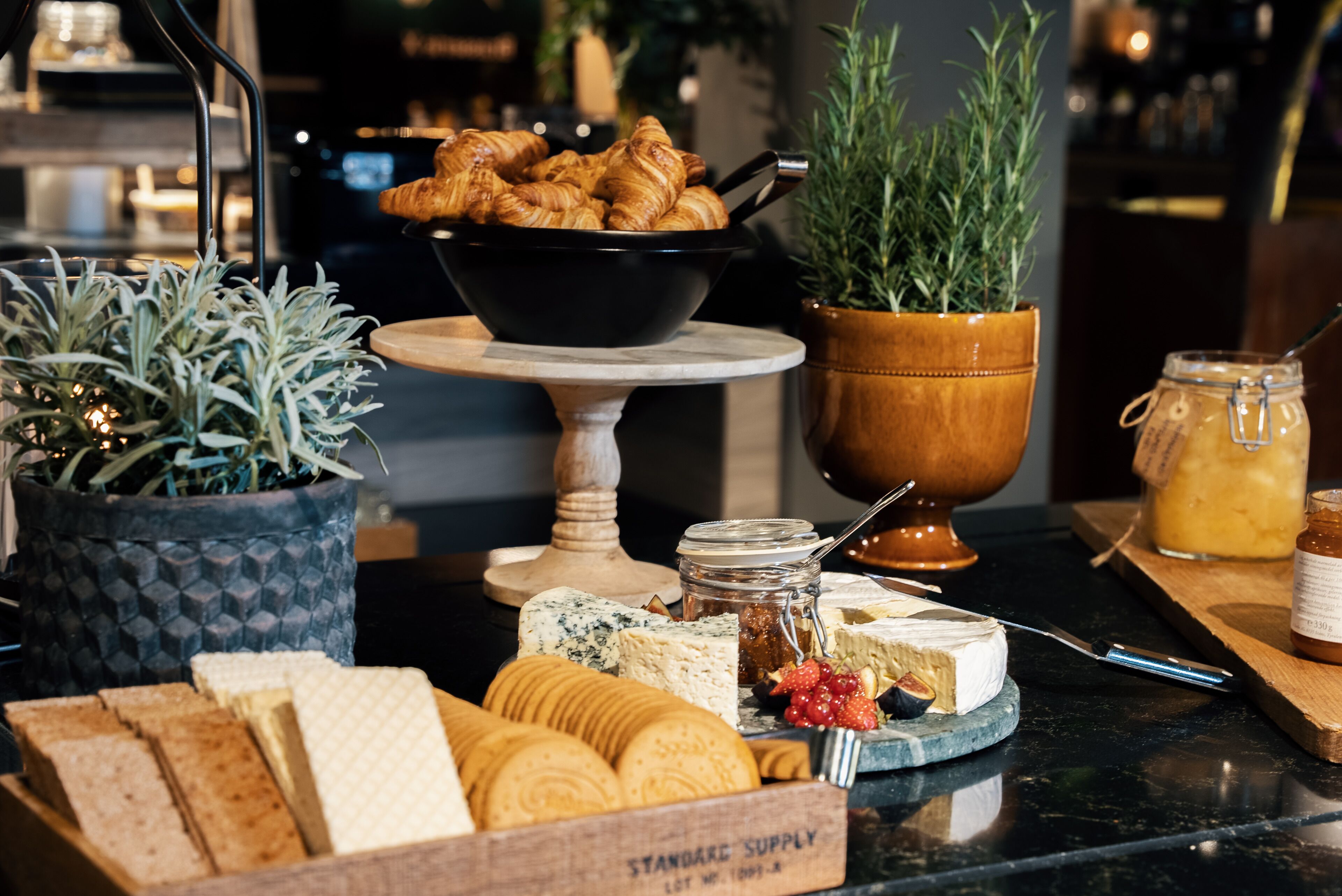 A variety of cheeses, crackers, and pastries on a marble countertop, with a potted plant and a bowl of croissants in the background.