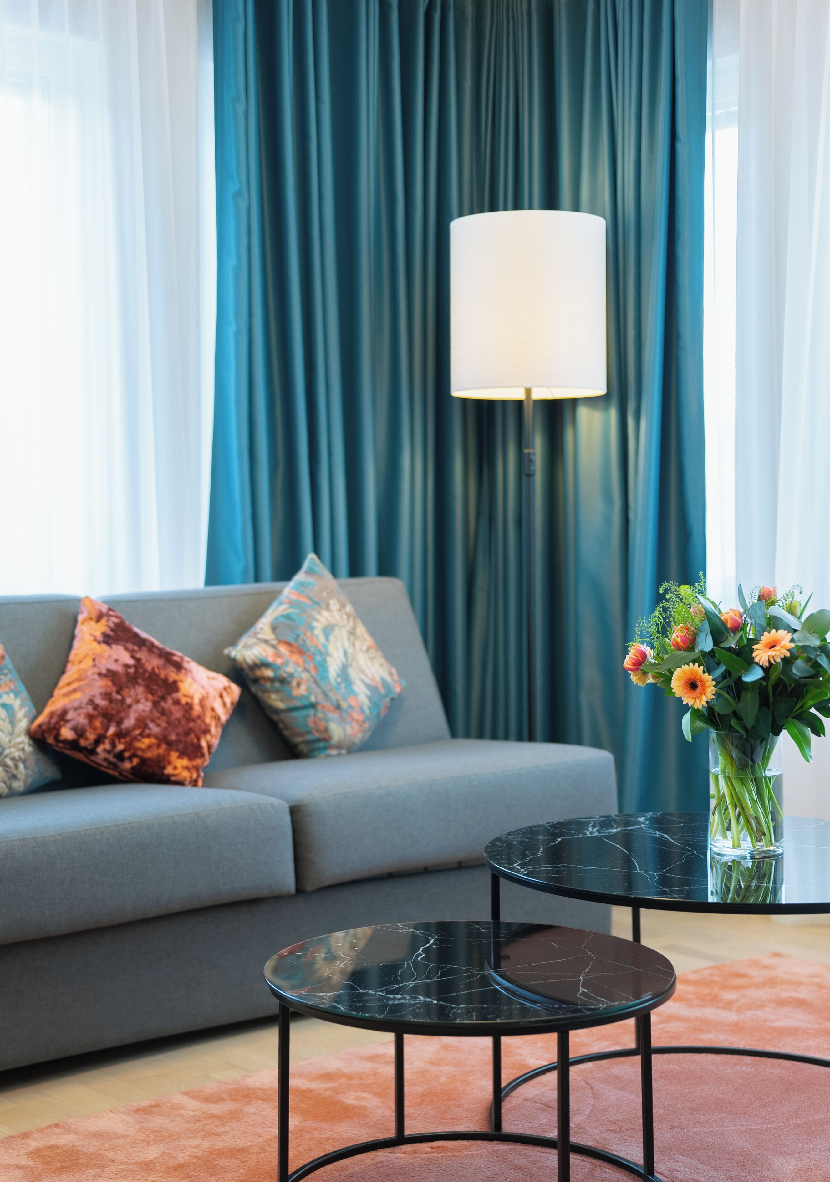 Un salon moderne avec un canapé gris, des coussins à motifs, une table basse en marbre noir et un vase rempli de fleurs oranges.