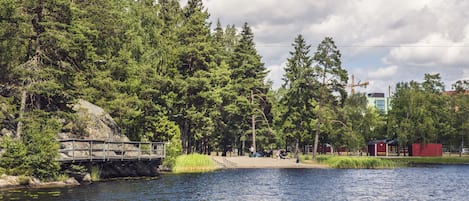 Un parque junto al lago con un puente de madera, zonas de picnic y edificios rojos al fondo.