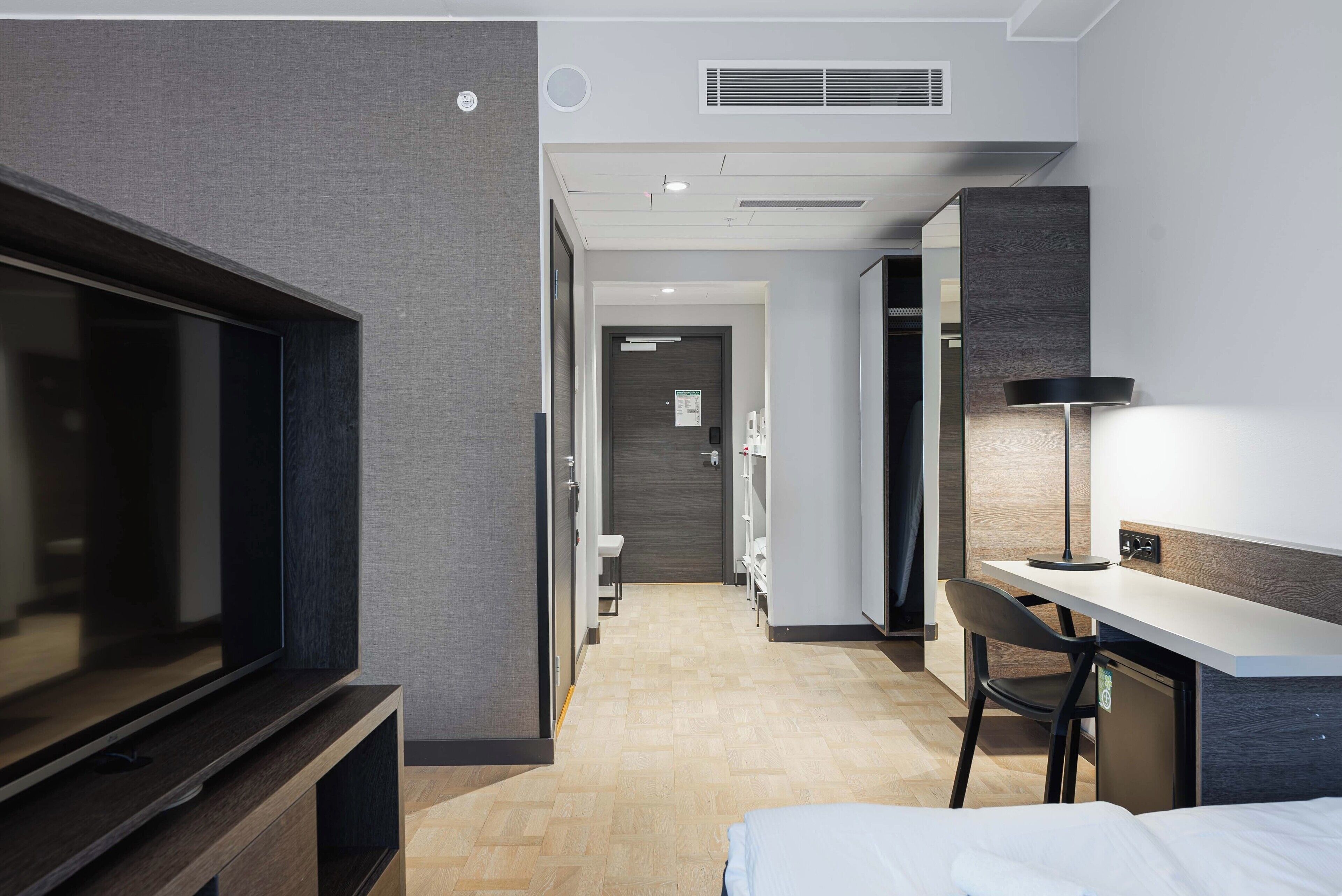 Una habitación de hotel moderna con una televisión de pantalla plana, un escritorio con lámpara, una cama y un armario.