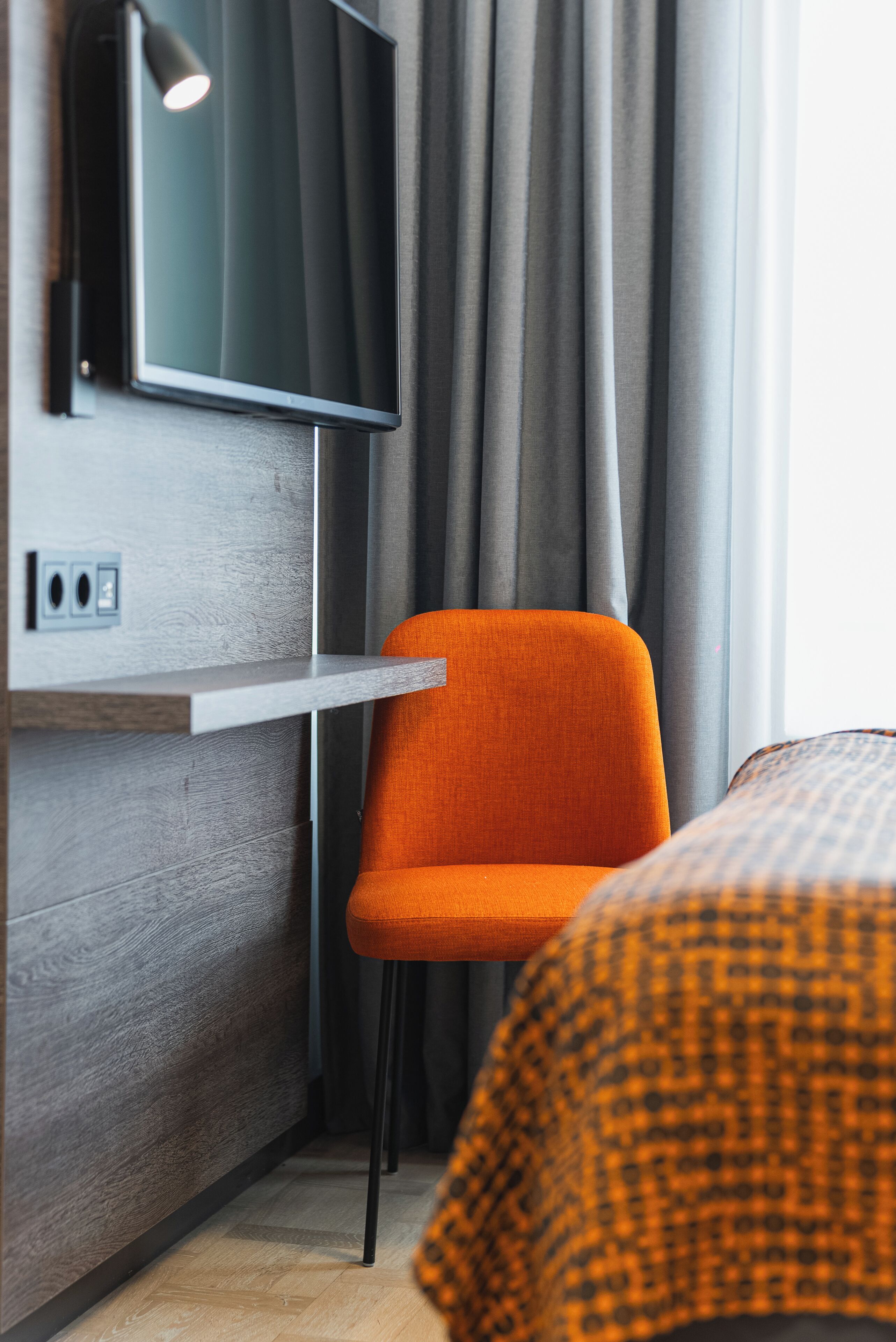 A hotel room with a modern design, featuring a flat-screen TV mounted on the wall, a wooden panel wall, a grey curtain, and an orange chair.