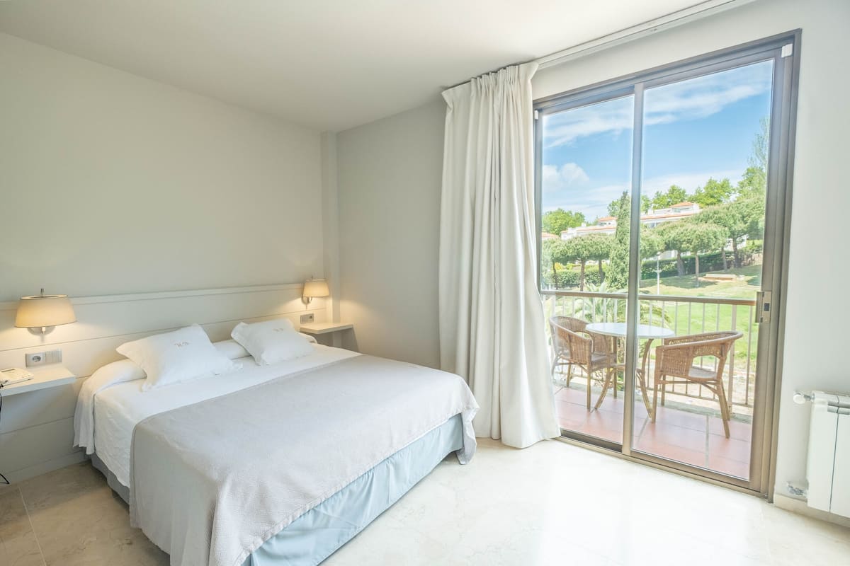 A hotel room with a bed, two bedside tables with lamps, a balcony with wicker chairs, and a view of greenery and buildings.