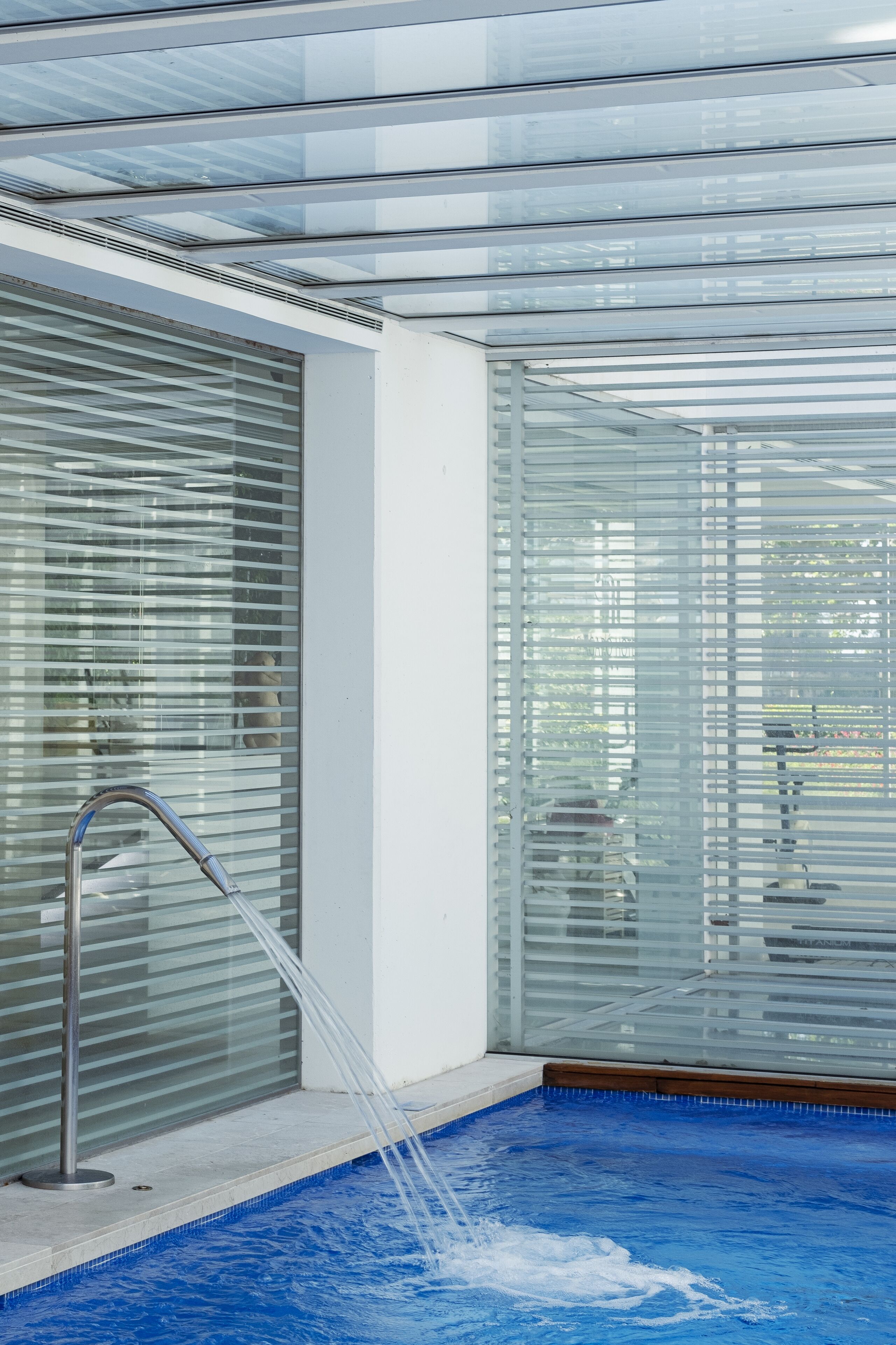 Un área de piscina interior moderna con paredes de vidrio transparente, un grifo metálico y suelo de color azul.