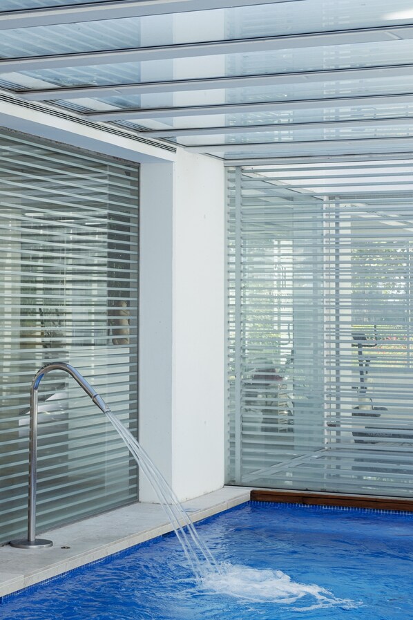 Un área de piscina interior moderna con paredes de vidrio transparente, un grifo metálico y suelo de color azul.
